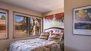 A bedroom with a large bed, two windows, a painting, and a framed picture on the wall.