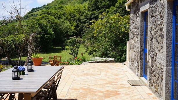 Un bâtiment en pierre avec des portes bleues, une terrasse avec une table et des chaises en bois, et une colline verdoyante en arrière-plan.