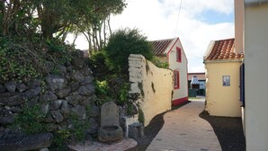 Un étroit sentier pavé de briques mène à un bâtiment doté d’une porte rouge, entouré de verdure et d’un mur de pierre.