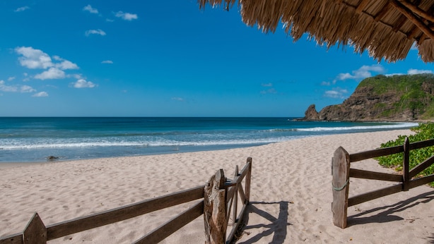 Una playa con una valla de madera y una estructura de techo de paja.