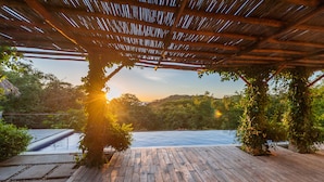 Una terraza de madera con pérgola, rodeada de abundante vegetación y vista a un cuerpo de agua.