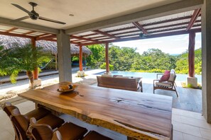 A wooden dining table with chairs, a ceiling fan, and a view of a swimming pool and lush greenery. - Exclusive Two Story Ocean View Home/private Pool ++privacy - Families Welcome! (San Juan del Sur)