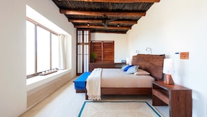 Un dormitorio con techo de madera, un ventanal grande, una cama con almohadas, una mesita de noche y una mesilla.