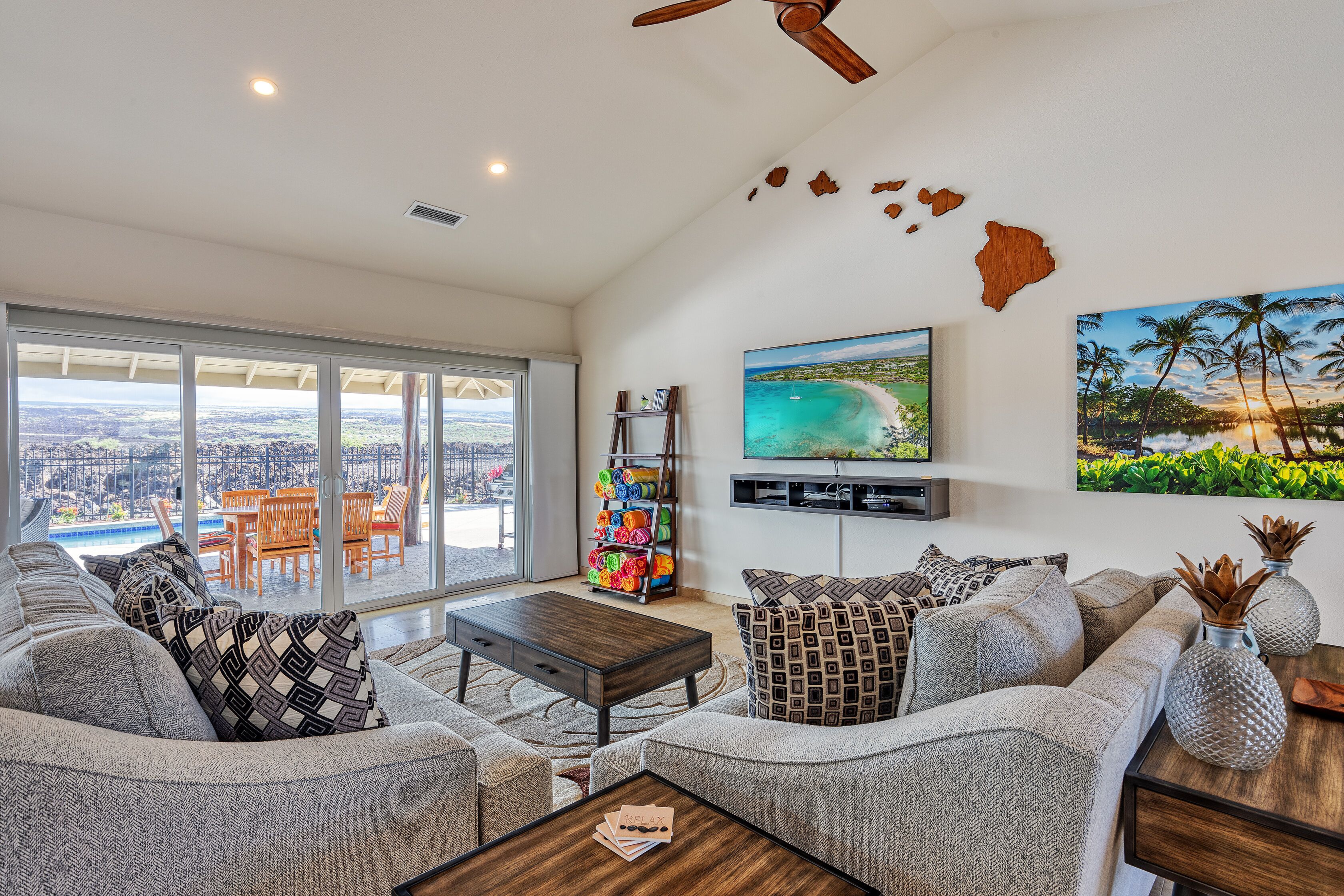 A spacious living area with a large window overlooking a cityscape, a flat-screen TV mounted on the wall, and a ceiling fan.
