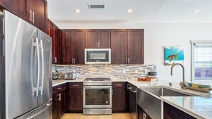 A modern kitchen with dark wood cabinets, stainless steel appliances, a tiled backsplash, and a large window with blinds. - 3 Bedroom Home with a Pool at Waikoloa Beach Resort (Waikoloa)