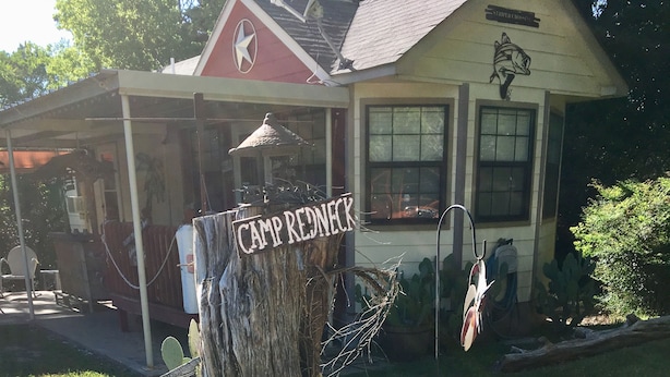 A rustic outdoor setting with a wooden sign reading "Camp Redneck," a bird bath, a metal pot, and a decorative stone slab. -  CLEAN, COZY LAKE CABIN COMFORT... (Clifton)