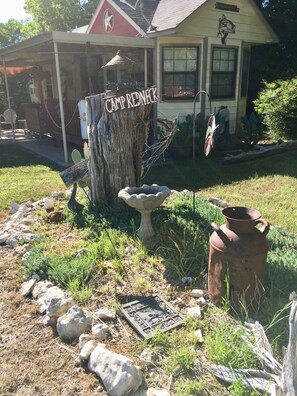 Ambiente rústico ao ar livre com uma placa de madeira que diz “Camp Redneck”, um banho para pássaros, uma panela de metal e uma laje de pedra decorativa.