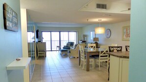 A spacious living area with a dining table, chairs, and a sofa. There is a staircase leading to an upper level, and a large window providing natural light.