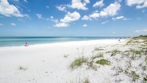 A beach with white sand, clear blue water, and sparse vegetation.