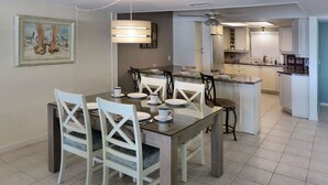A dining area with a glass table, white chairs, and a kitchen in the background.
