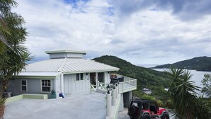 A building with a covered entrance, a balcony, and a view of a body of water and hills. - Million Dollar Panoramic Views! Huge Decks and  Pool! (Coral Bay)