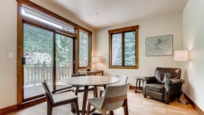 A room with a wooden floor, a round table with chairs, a leather armchair, and large windows with a view of trees.