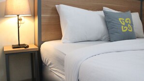 Un lit bien fait, avec une literie blanche et deux oreillers : un uni, blanc, et l’autre orné d’un logo jaune. Une lampe de chevet est posée sur une table de nuit en bois, et le mur est agrémenté d’une tête de lit en bois.