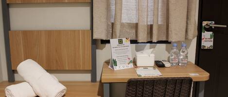 A hotel room with a wooden headboard, a desk with a remote control, two Le Minetale water bottles, a tissue box, and a sign indicating check-in time.