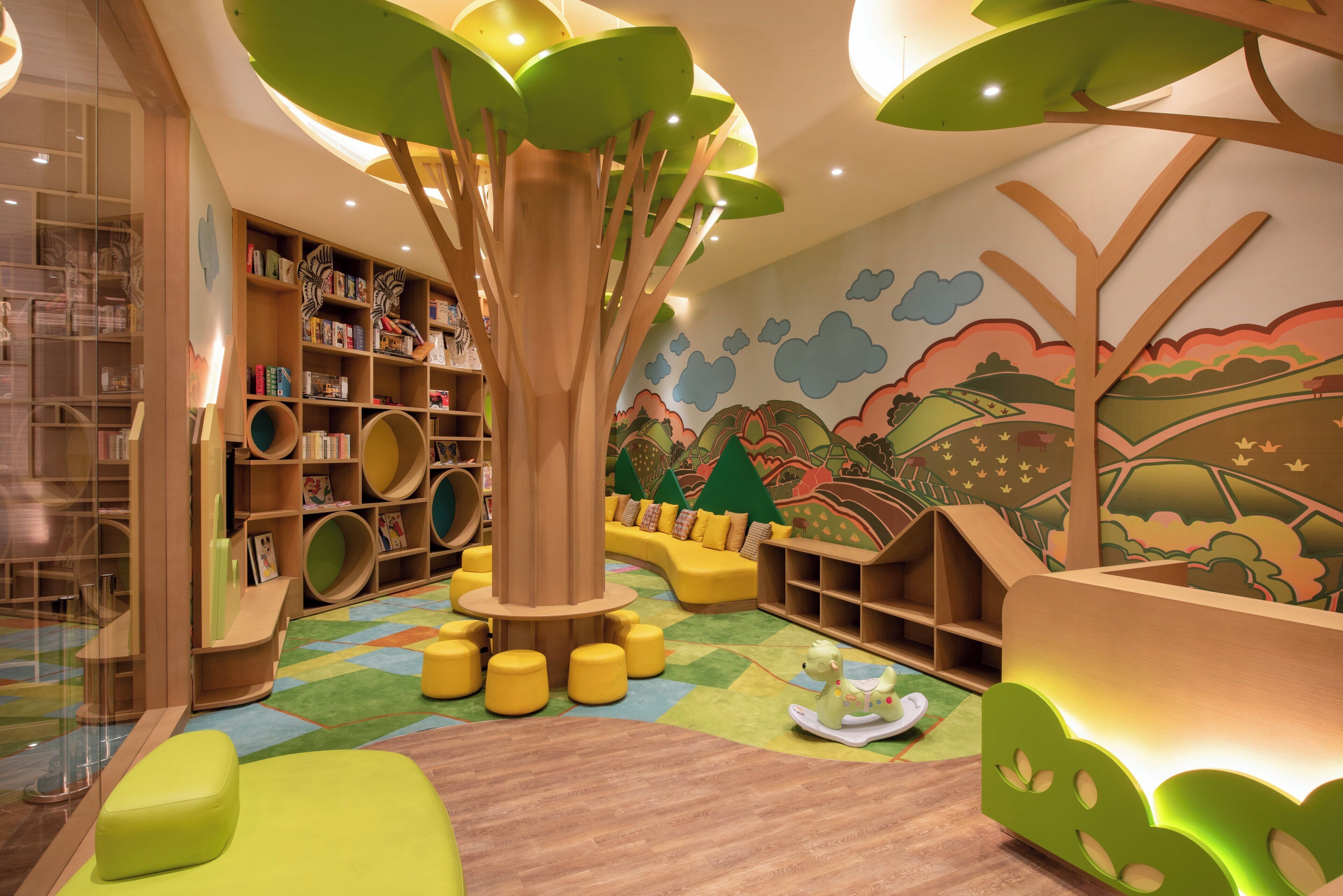 A children's play area with a large tree structure, colorful bookshelves, and a mural on the wall.
