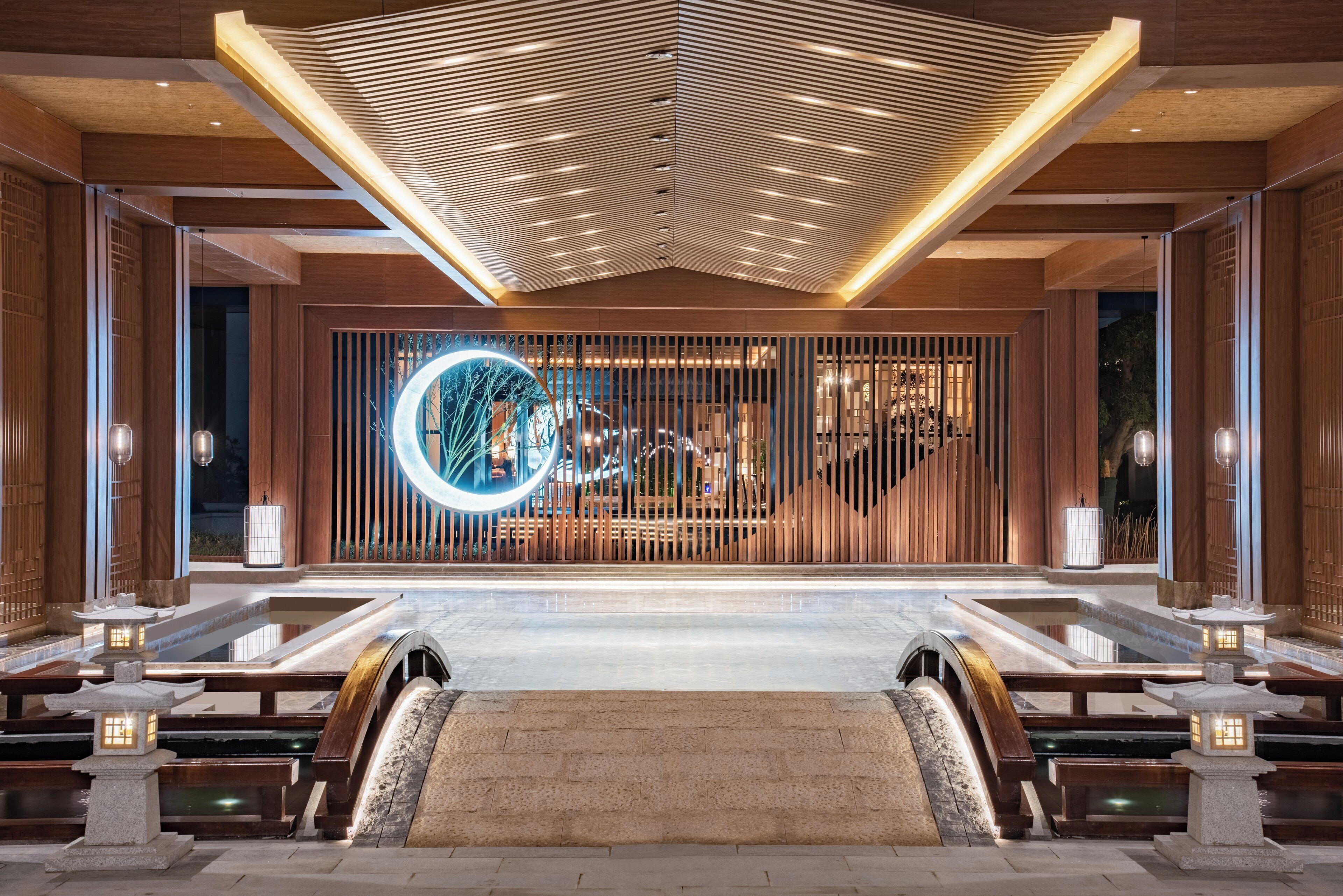 A modern hotel lobby with a central reception desk, a decorative wooden panel wall, and a circular illuminated sign.