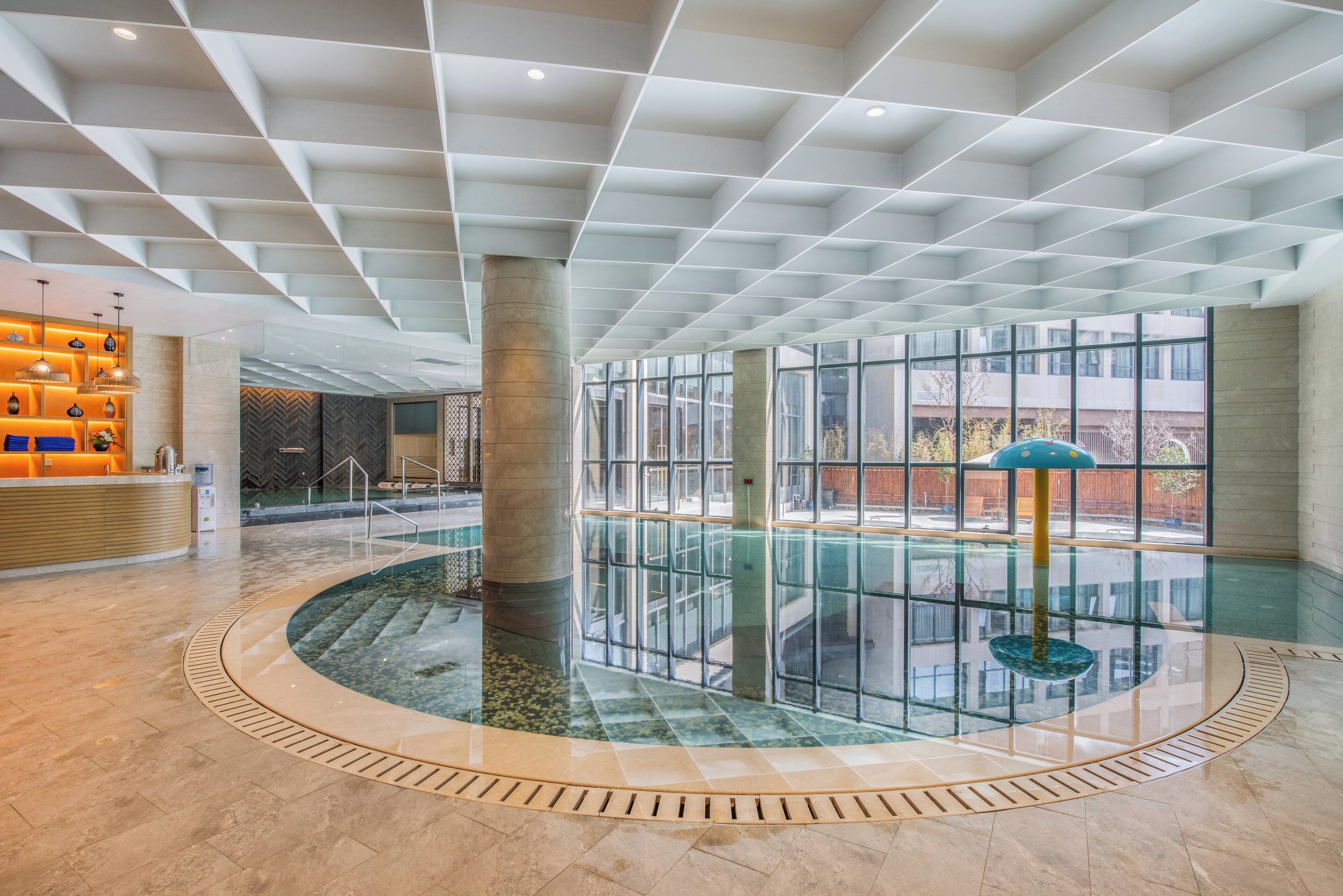 A spacious indoor swimming pool with a glass wall, surrounded by a tiled floor, and a reception area with a wooden counter and decorative lighting.