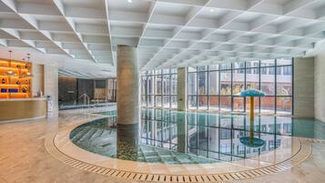 A spacious indoor swimming pool with a glass wall, surrounded by a tiled floor, and a reception area with a wooden counter and decorative lighting.