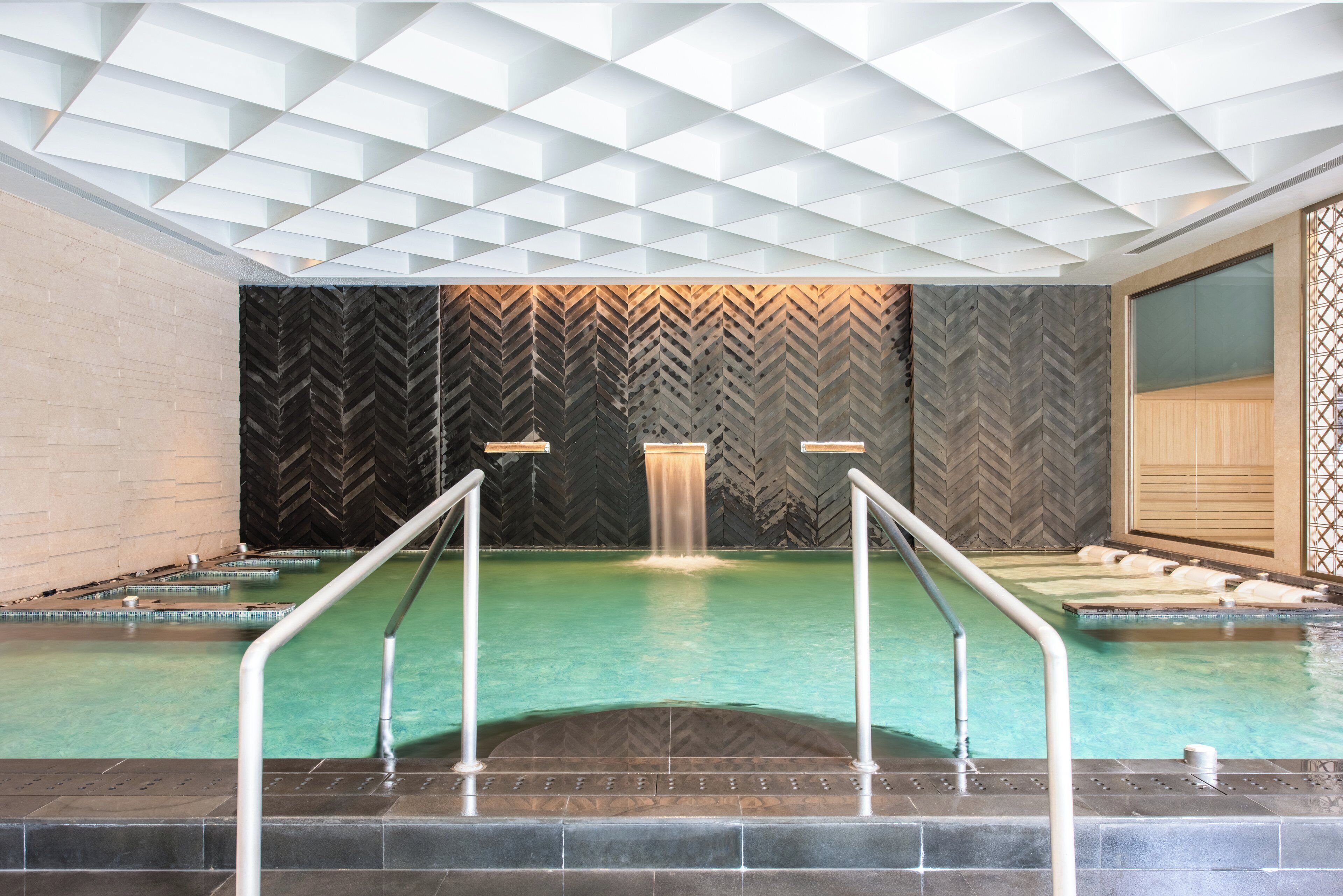 A modern indoor pool with a geometric ceiling design, a central water feature, and a herringbone-patterned wall.
