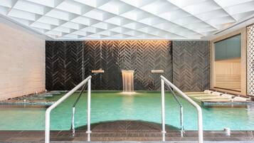 A modern indoor pool with a geometric ceiling design, a central water feature, and a herringbone-patterned wall.