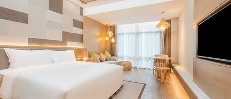 Una habitación de hotel moderna con una cama grande, televisión, zona de estar con una mesa y sillas, y un amplio ventanal con cortinas.