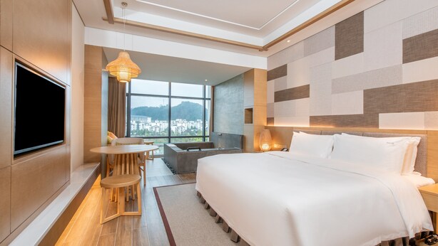 A modern hotel room with a large bed, a TV mounted on the wall, a dining area with a table and chairs, and a view of the city through large windows. - Holiday Inn Resort Yichun Mingyue Mountain by IHG (Yichun)