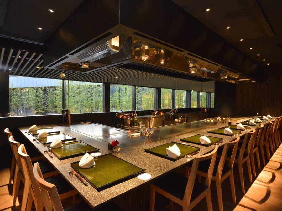 Une longue table à manger entourée de nombreuses chaises, un îlot central et une vue sur la verdure à l’extérieur.