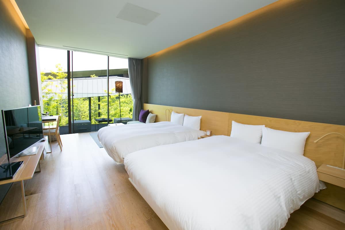 A modern hotel room with two beds, a desk with a flat-screen TV, and a large window offering a view of greenery.