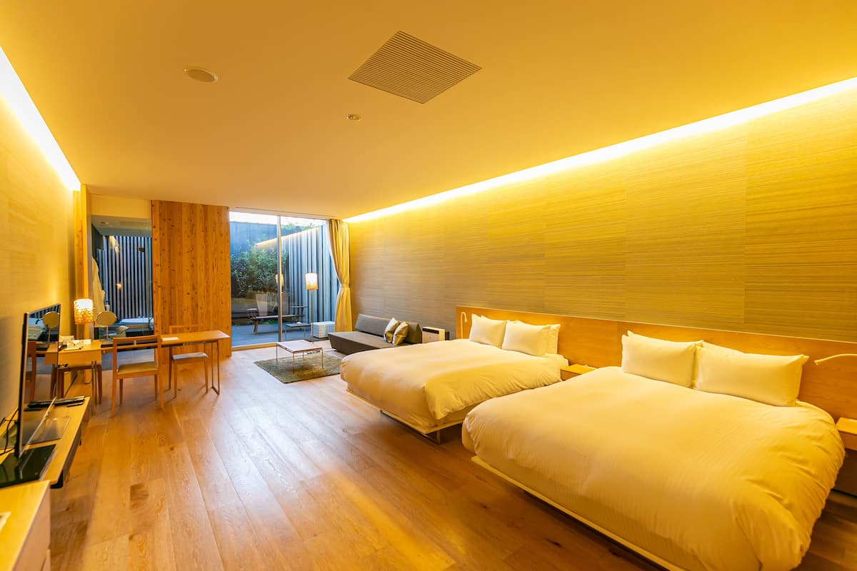A modern hotel room with two beds, a desk, and a seating area with a view of the outdoors.