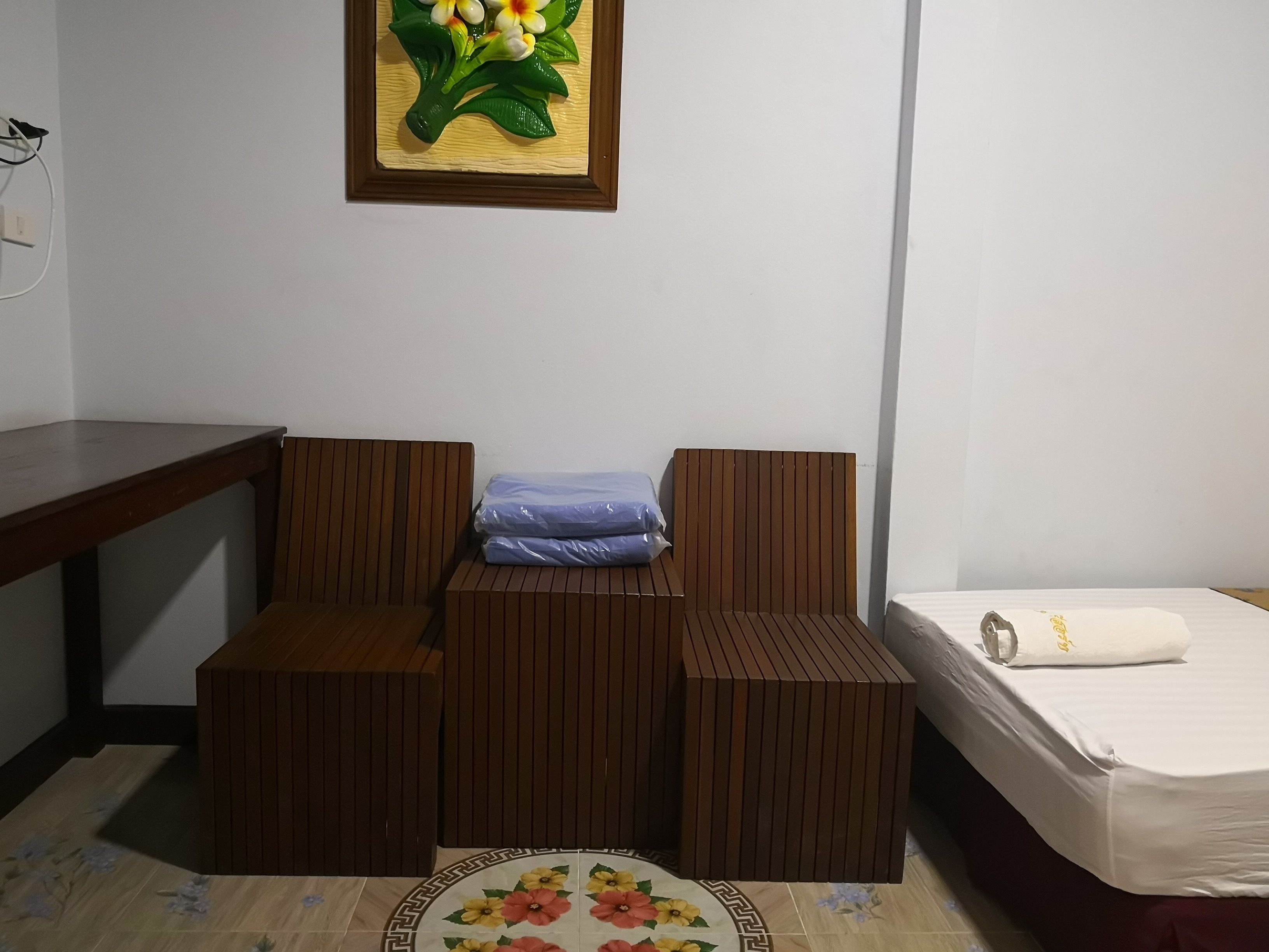 A small, neatly arranged room with a wooden desk, a cushioned seating area, a bed with a white mattress and a pillow, and a floral-patterned rug.