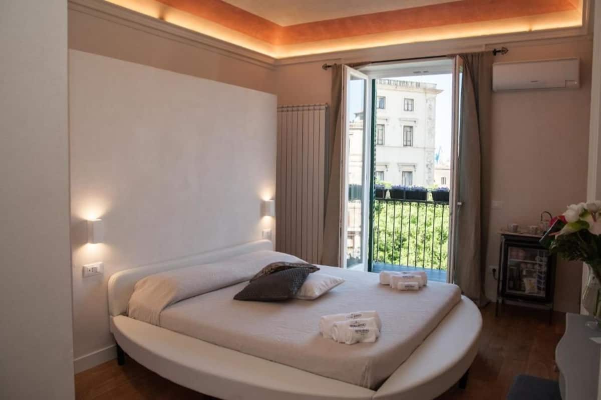 Een moderne slaapkamer met een rond bed, een balkon met uitzicht op gebouwen, en een tafeltje met een bloevas.