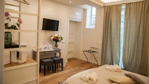 Una habitación de hotel con una cama, un escritorio con una silla, un televisor, una ventana con cortinas y una pequeña mesa con un jarrón de flores.