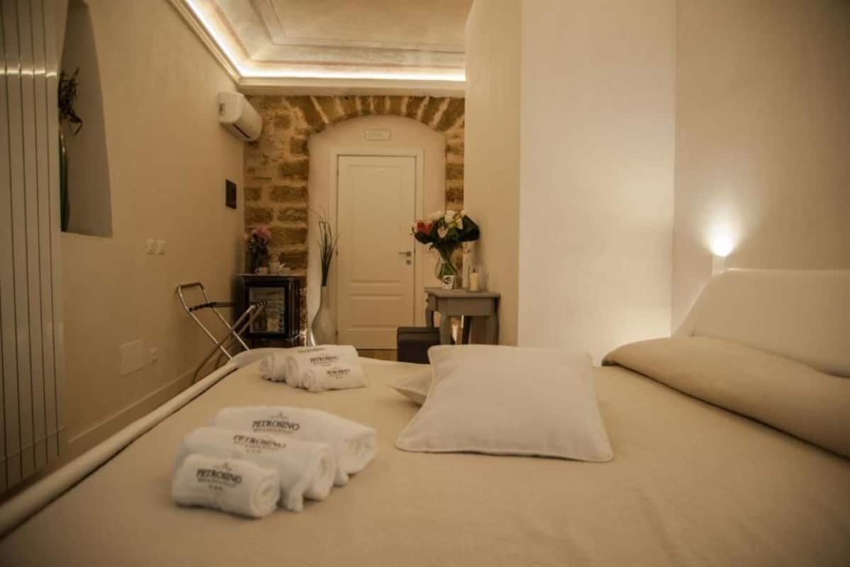 Een hotelkamer met een bed, een kleintje tafeltje met een bloemetjesvaas, een handdoekhouder en een stenen muur.