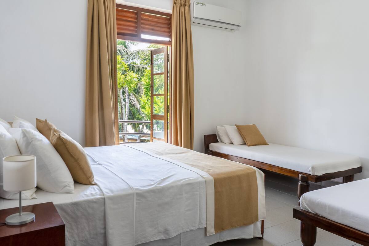 A hotel room with a large bed, a smaller bed, a nightstand with a lamp, a window with curtains, and an air conditioning unit.
