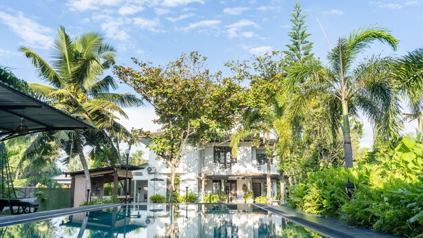 A swimming pool surrounded by lush greenery and a white building with a tiled roof. - Villa Sarakkuwa (Negombo)