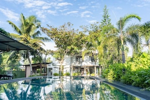 A swimming pool surrounded by lush greenery and a white building with a tiled roof.