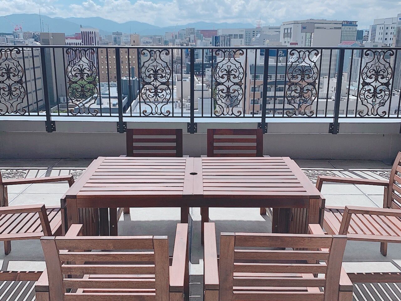 Una terraza en la azotea con mesa y sillas de madera, barandilla metálica decorativa y vista panorámica de la ciudad.