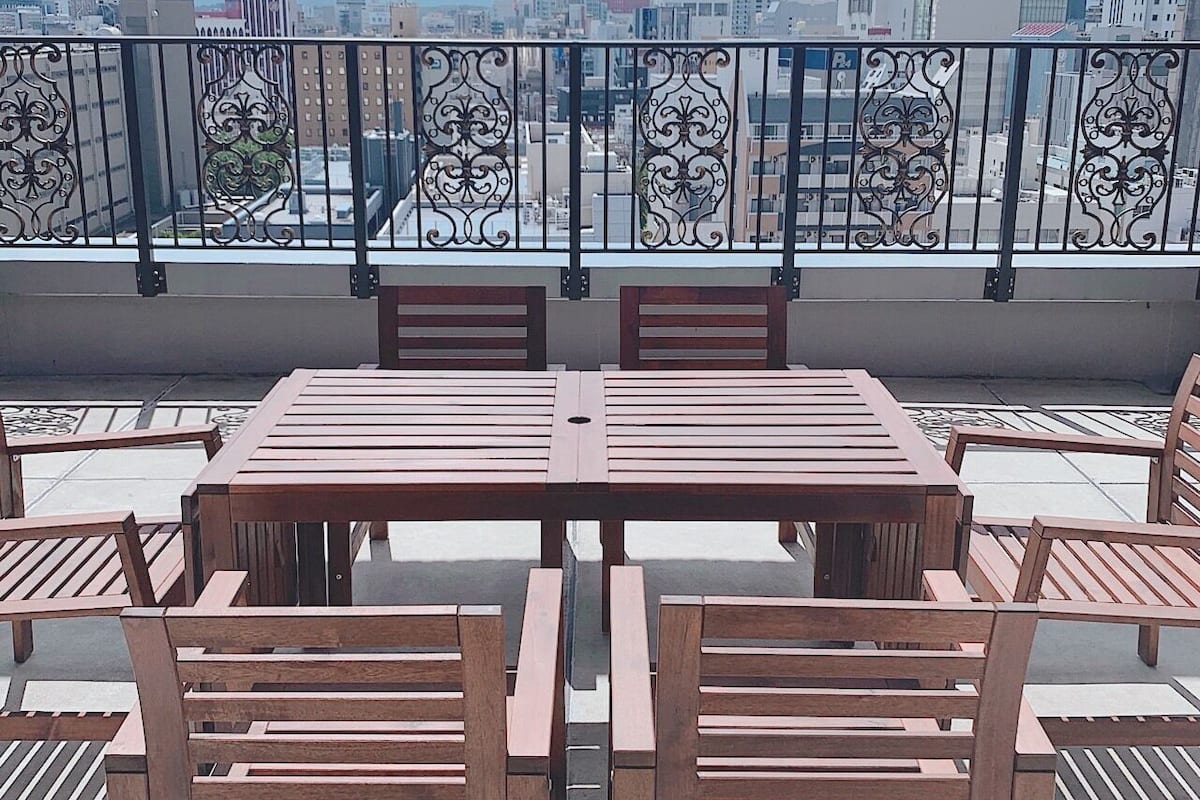 A rooftop terrace with a wooden table and chairs, a decorative metal railing, and a cityscape view.