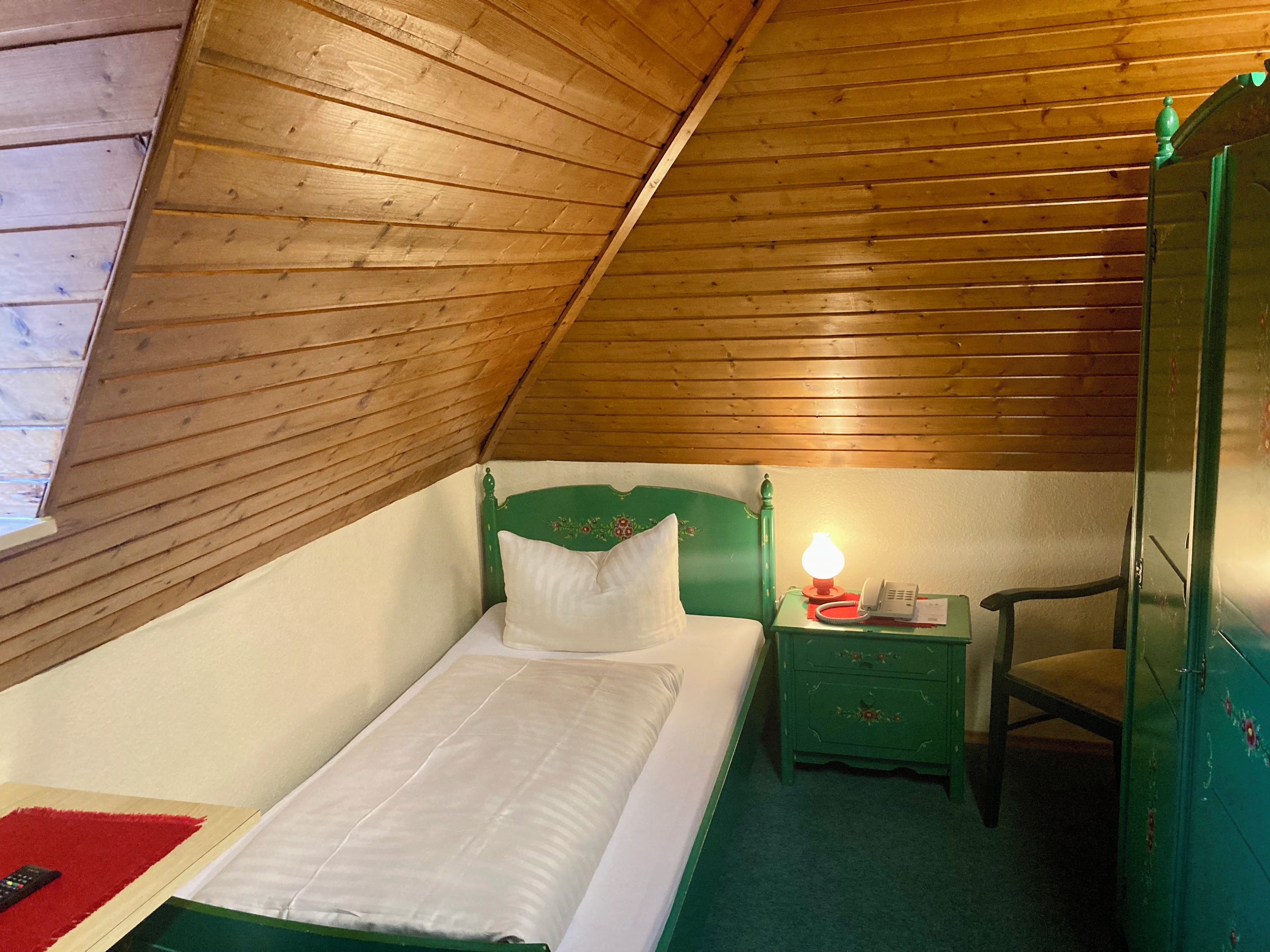 A single bed with a green headboard, a bedside table with a lamp, a chair, and a wardrobe in a room with wooden sloped ceilings.