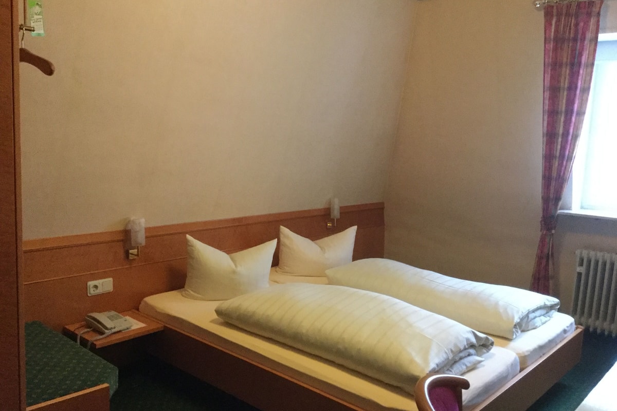 A hotel room with a double bed, a bedside table with a phone, a green upholstered bench, and a window with curtains.