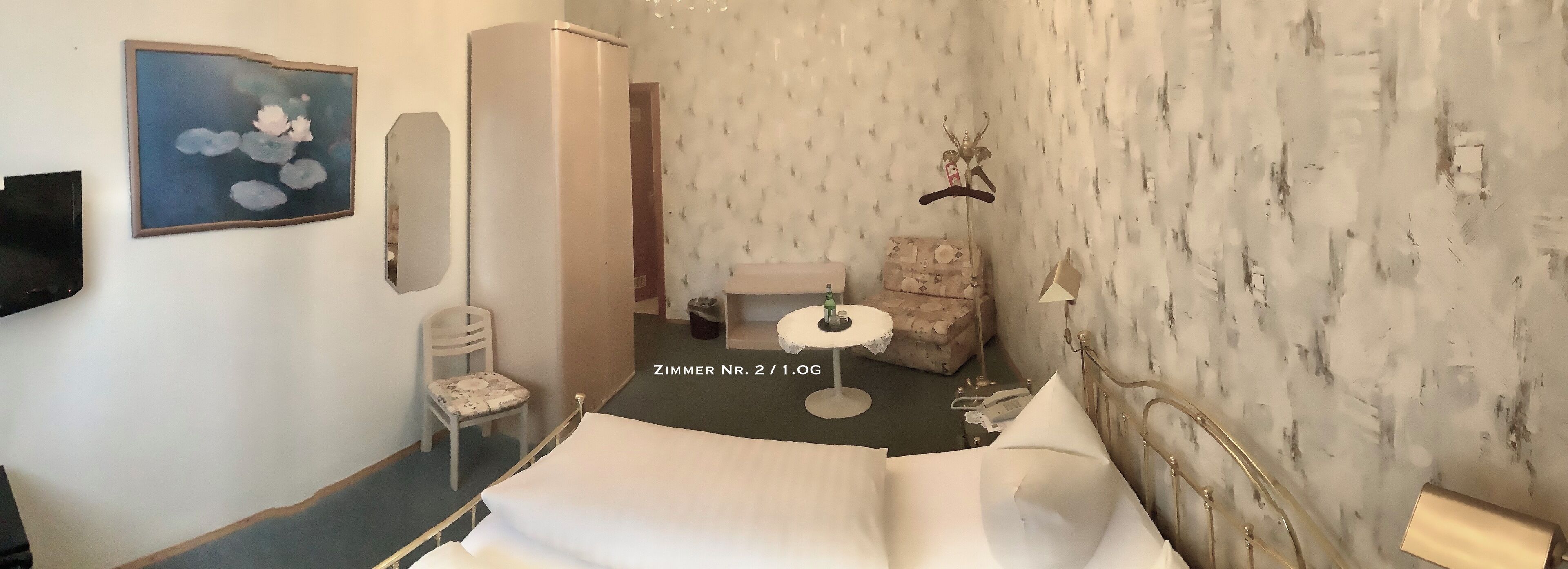 A hotel room with a bed, a chair, a small table, a painting, and a wardrobe.