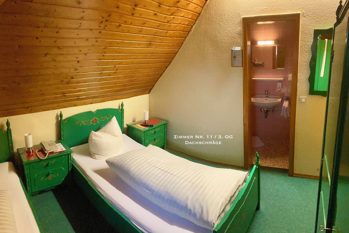 A room with a sloped ceiling, green furniture, a bed with white bedding, a nightstand with a lamp, and a green-framed mirror.