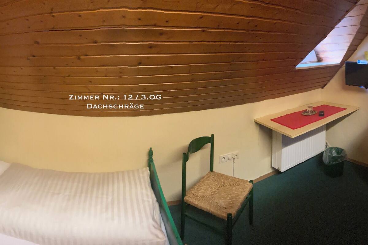 A small, cozy room with a bed, a chair, a small table with a red mat, and a television mounted on the wall.