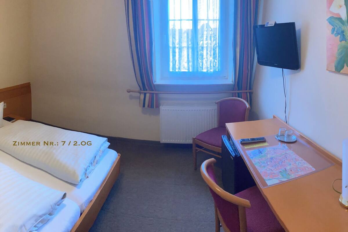 A hotel room with a double bed, a desk with a chair, a telephone, a radiator, and a window with curtains.