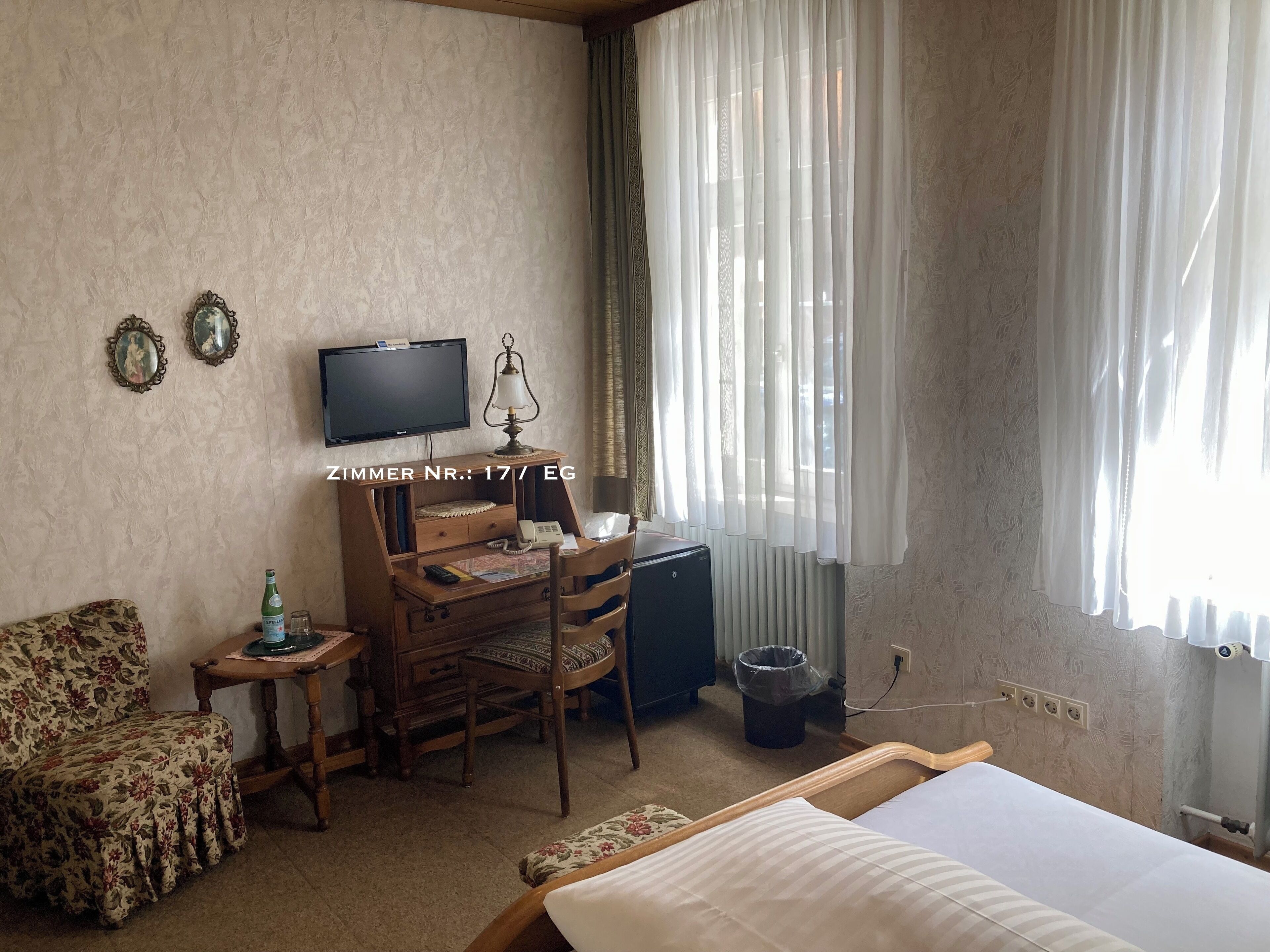 Quarto de hotel com cama, escrivaninha com luminária, cadeira, televisão e janela com cortinas.