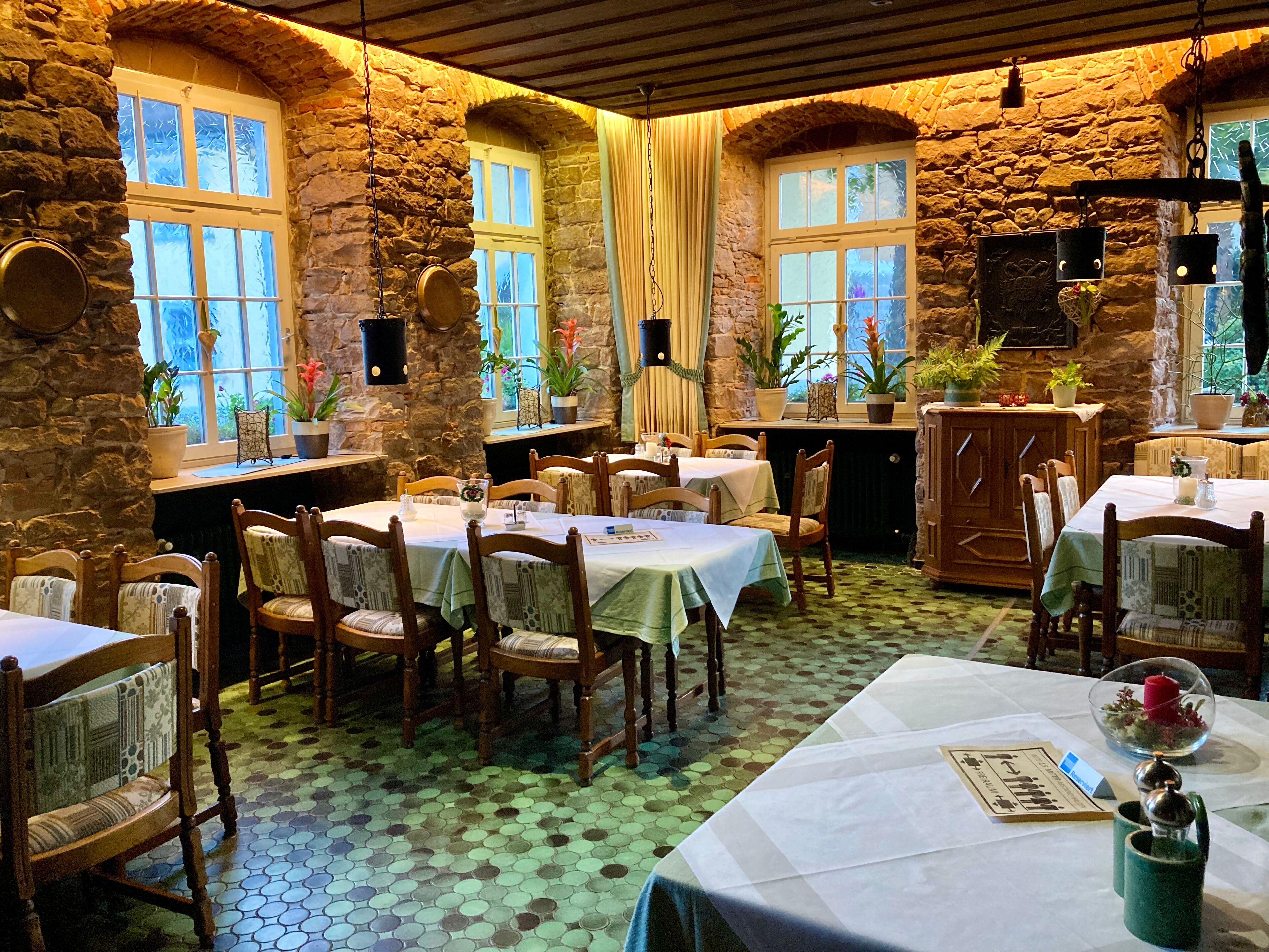 A rustic dining area with stone walls, multiple tables, and chairs, featuring a tiled floor and natural light from windows.