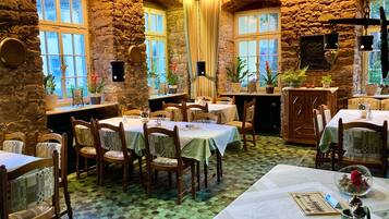 A rustic dining area with stone walls, multiple tables, and chairs, featuring a tiled floor and natural light from windows.