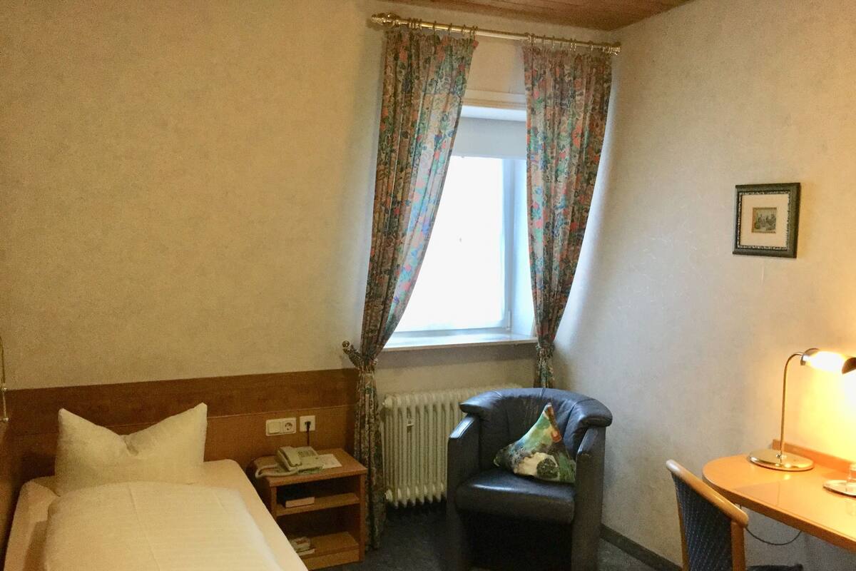 A hotel room with a bed, a chair, a desk with a lamp, a window with curtains, and a radiator.
