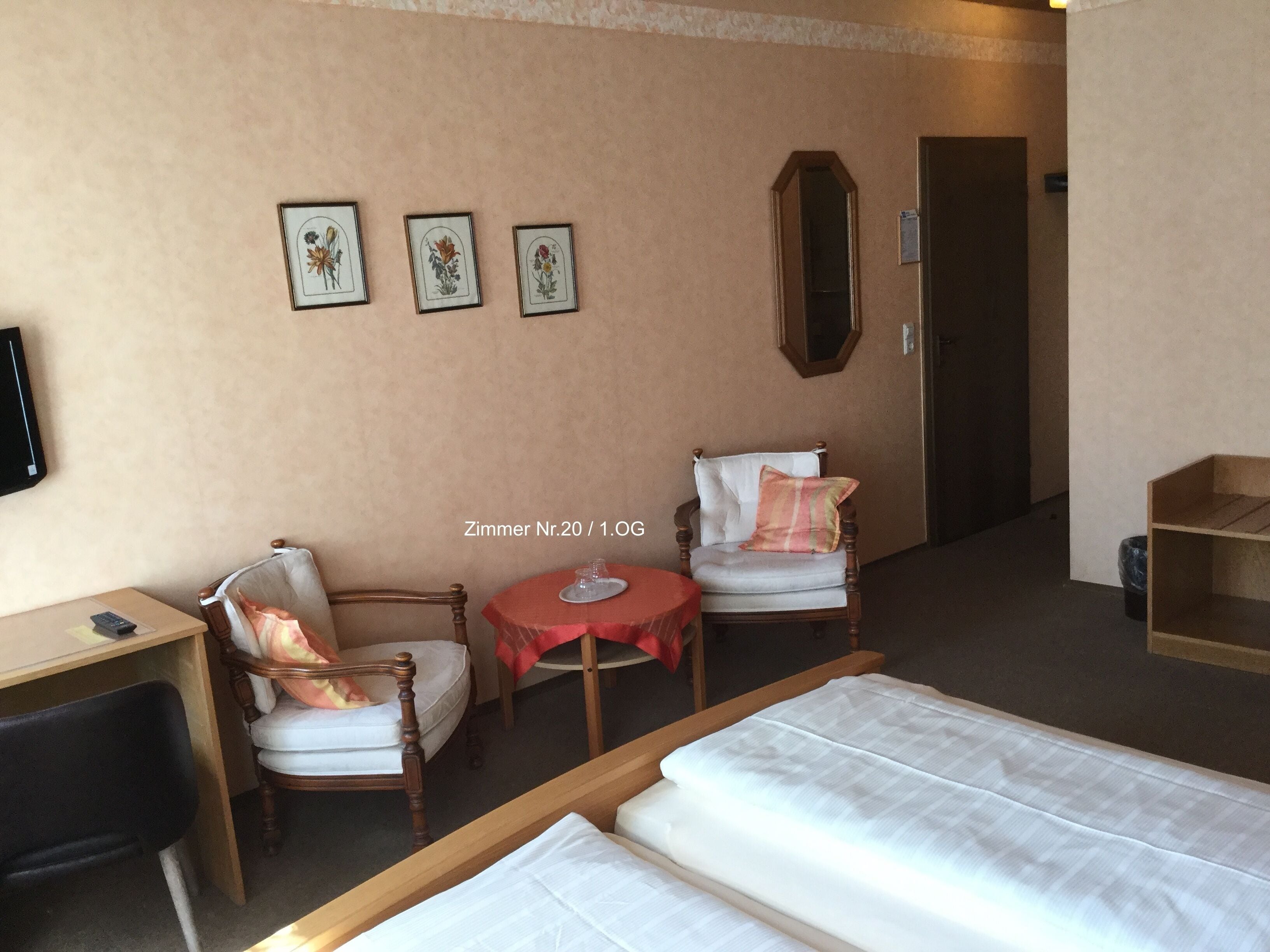 Habitación de hotel con dos camas individuales, un escritorio, una silla, una mesita con un vaso, y tres cuadros con motivos florales en la pared.