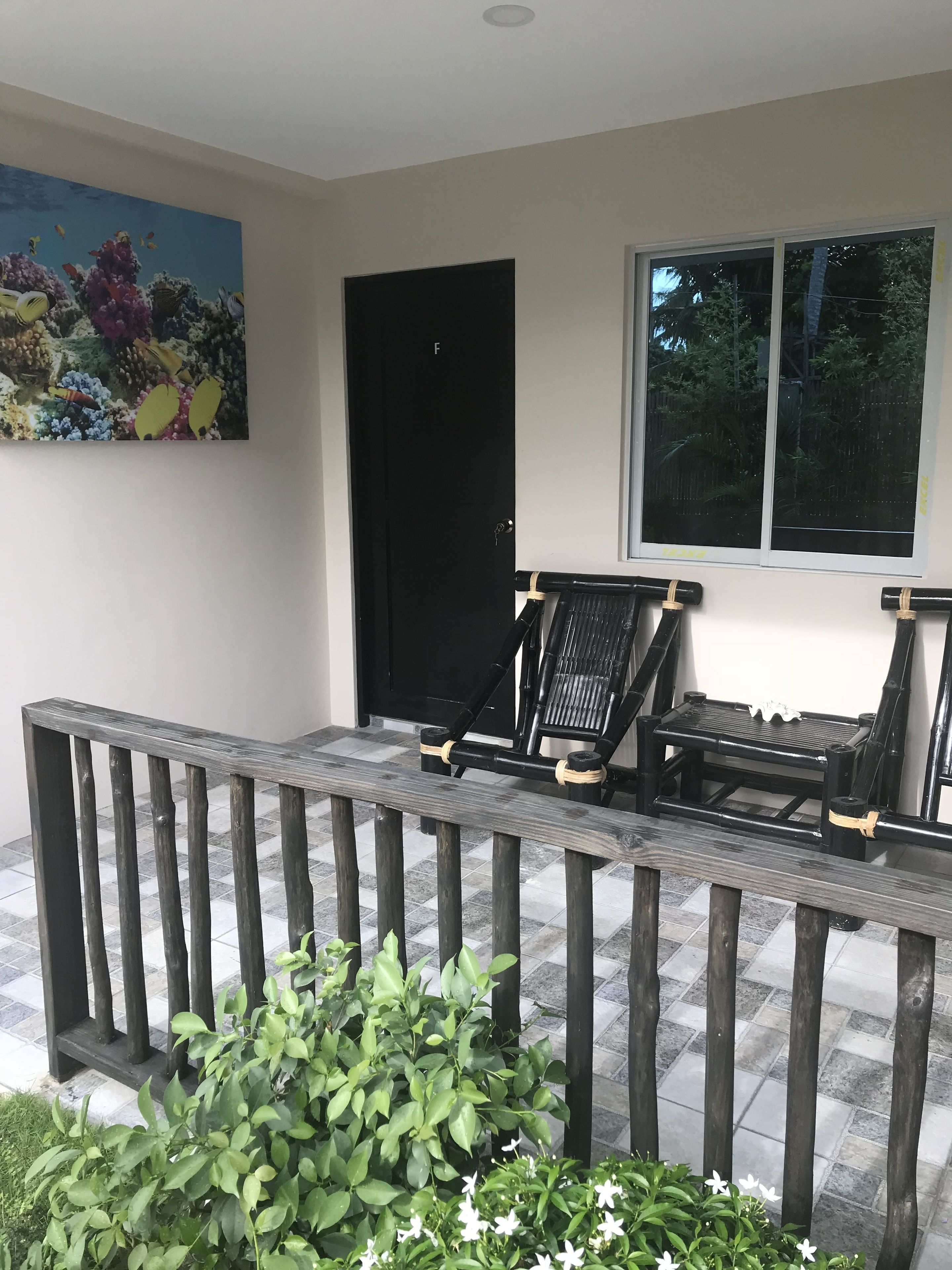 A covered outdoor seating area with two chairs and a table, a black door, a window, and a painting on the wall.
